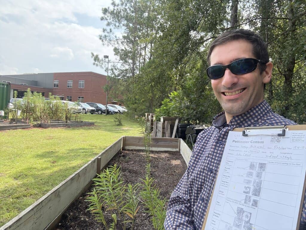 Man with sunglasses holding a clipboard in a garden with herbs and trees.