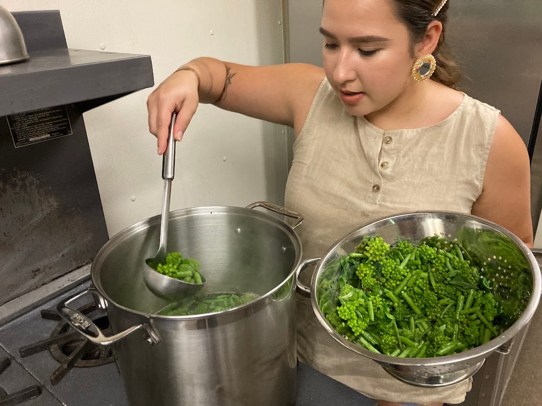 A woman ladling green milkweed buds into a stainless steel colander.