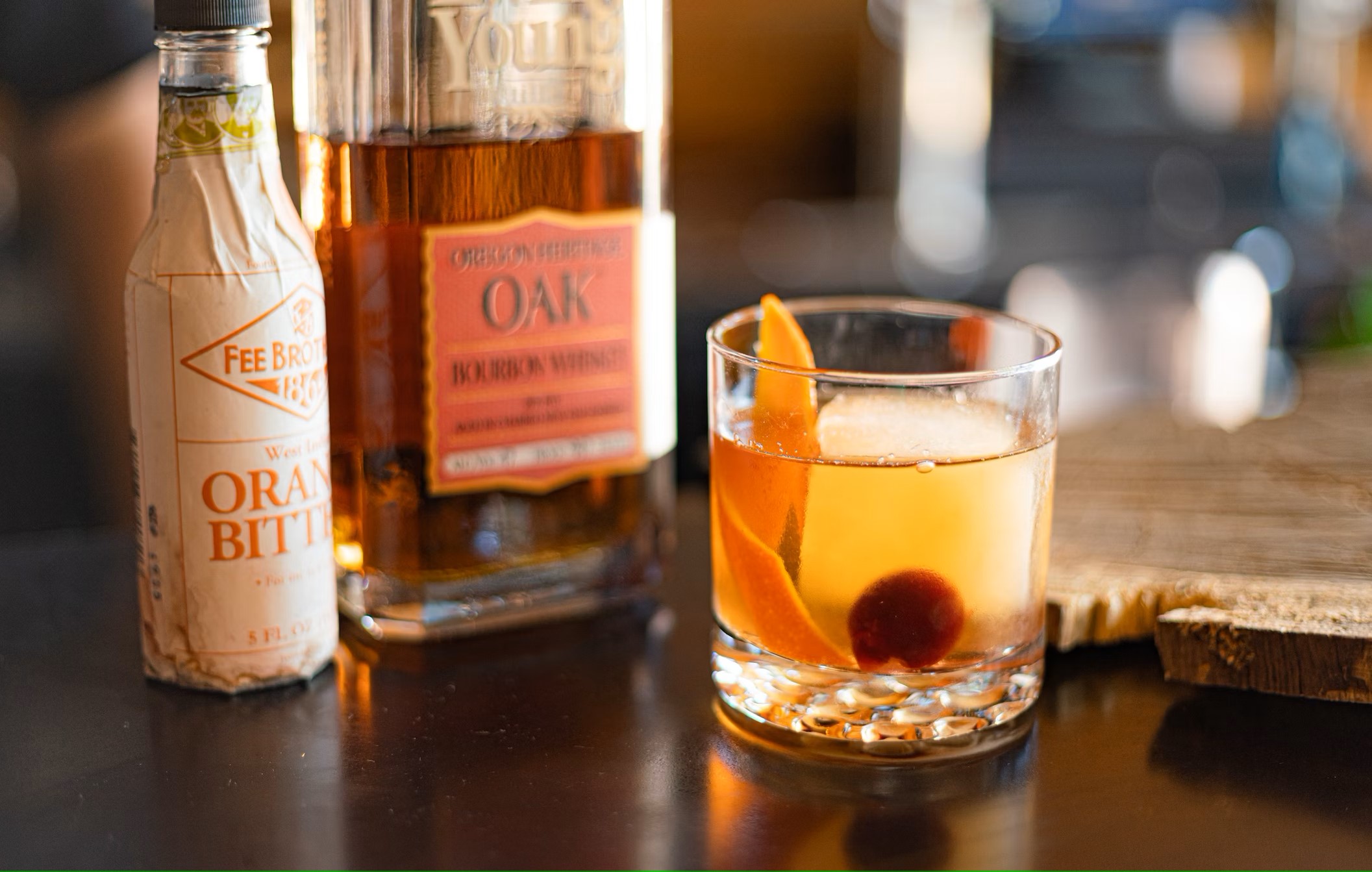 A yellow drink with a cherry and orange peel set on a bar with a bottle of bitters and one of liqour