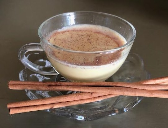 A clear glass tea cup with golden milk, sitting on a clear saucer with two cinnamon sticks